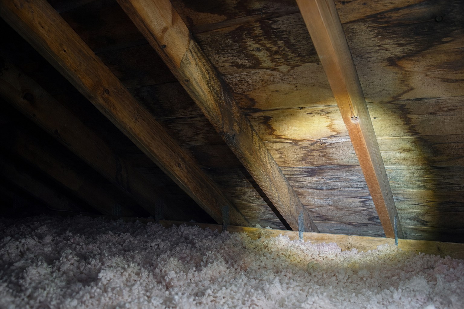 Entretoit d'une maison québécoise éclairé à la lampe, traces d'humidité visibles sur le platelage et les chevrons.
