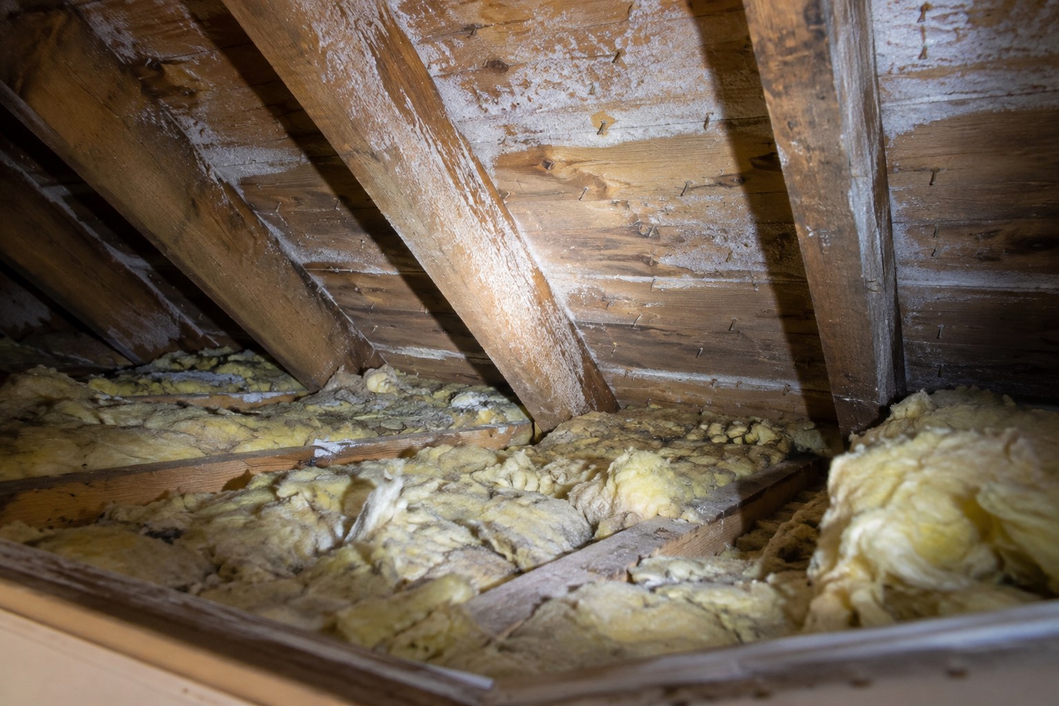 Entretoit vu depuis la trappe d’accès : fibre de verre jaunie, mince et inégale, et givre blanc sur les clous et le support de couverture.