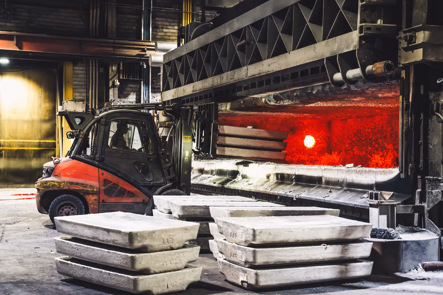 Forklift operating inside a factory near a glowing red-hot furnace with stacked metal molds on the floor.