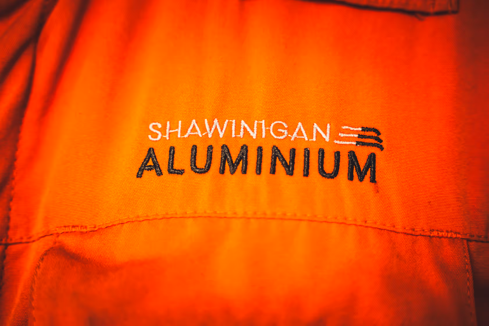 Close-up of orange fabric with embroidered text 'SHAWINIGAN ALUMINIUM' and a small logo above the text.