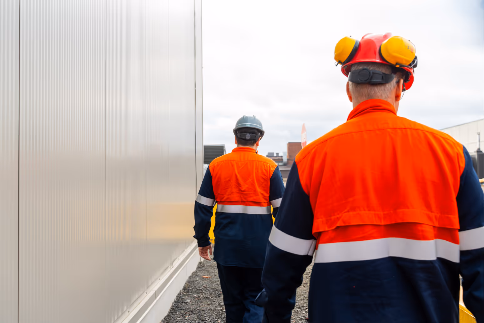 Two construction workers in orange and navy safety uniforms walking away beside a building wall, one wearing a red helmet with yellow ear protection.