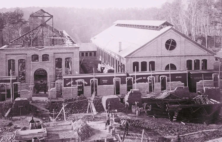 Black and white photo of an industrial railway building under construction with workers and horses in the foreground.