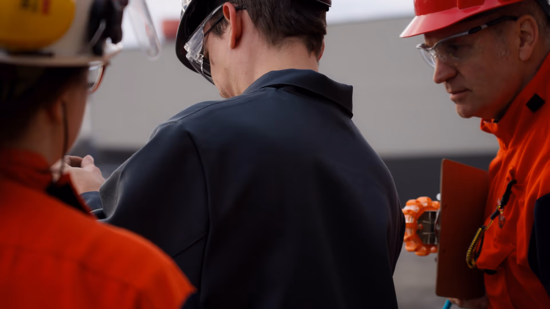 Three safety workers wearing helmets and protective glasses engaged in a discussion at a worksite.