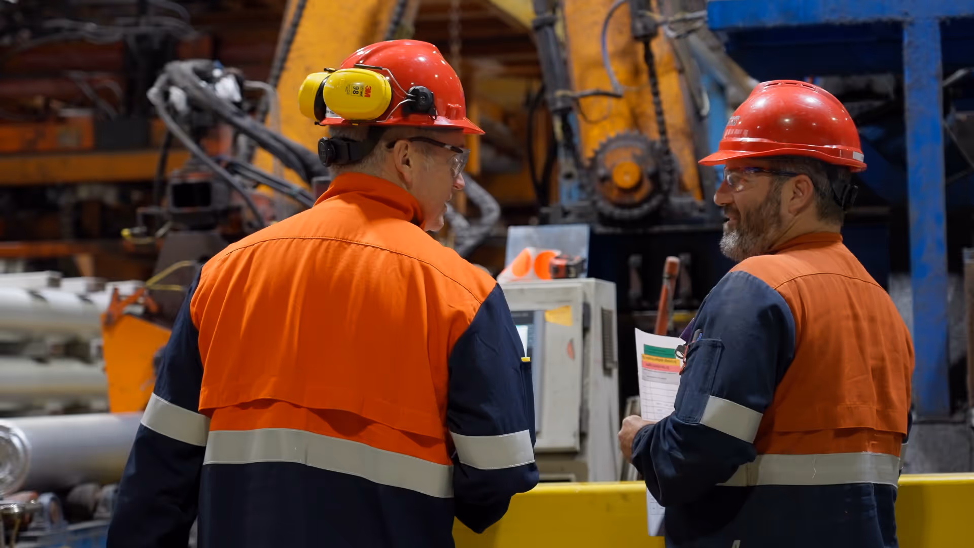 Two industrial workers wearing red hard hats and orange and navy protective clothing talking inside a factory setting.