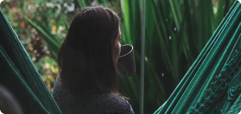 Daniella sitting in a hammock, facing away, drinking from a mug surrounded by green plants.