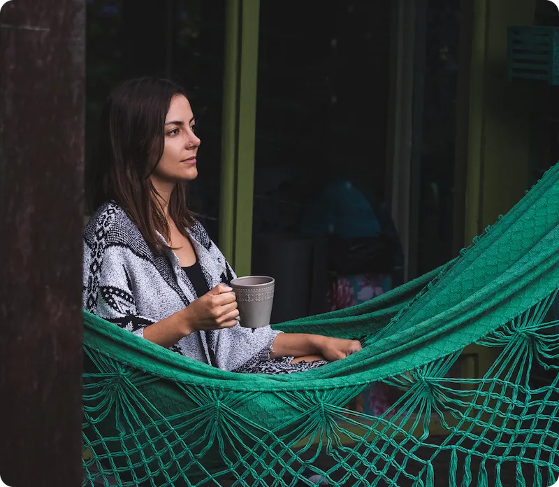 Daniella sitting in a green hammock holding a gray mug, wrapped in a patterned shawl and looking contemplatively to the left.