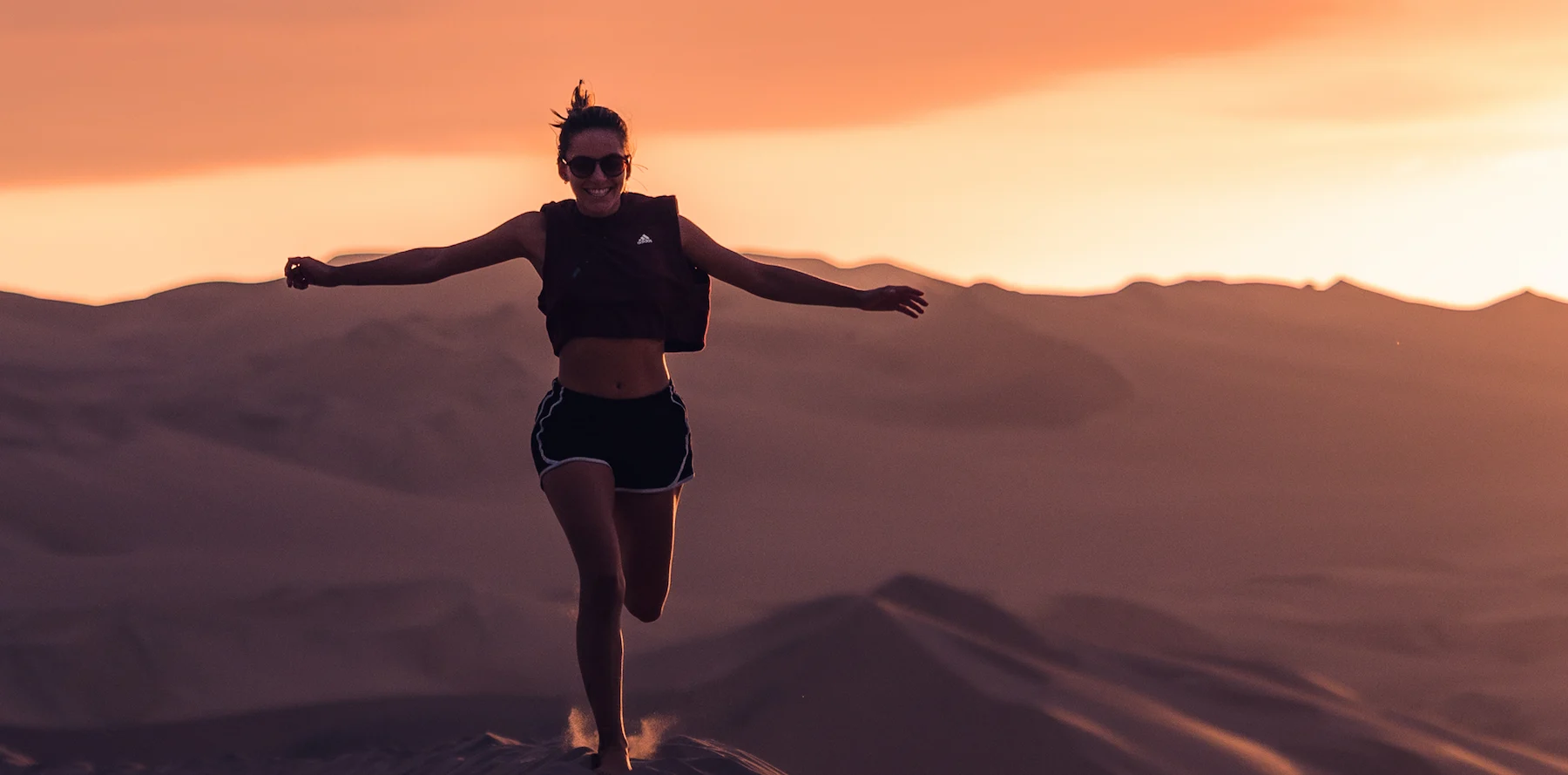 Happy woman running on sand dunes at sunset with arms outstretched and wearing sunglasses.