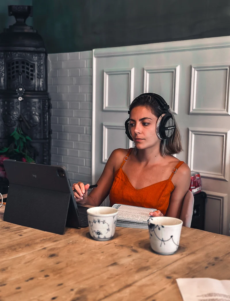 Woman wearing headphones and an orange sleeveless top working on a tablet with a notebook and pen at a wooden table with two ceramic cups.