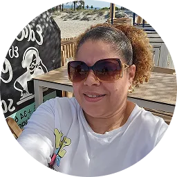 Woman wearing large sunglasses and a white shirt sitting outdoors at a wooden table with beach scenery in the background.
