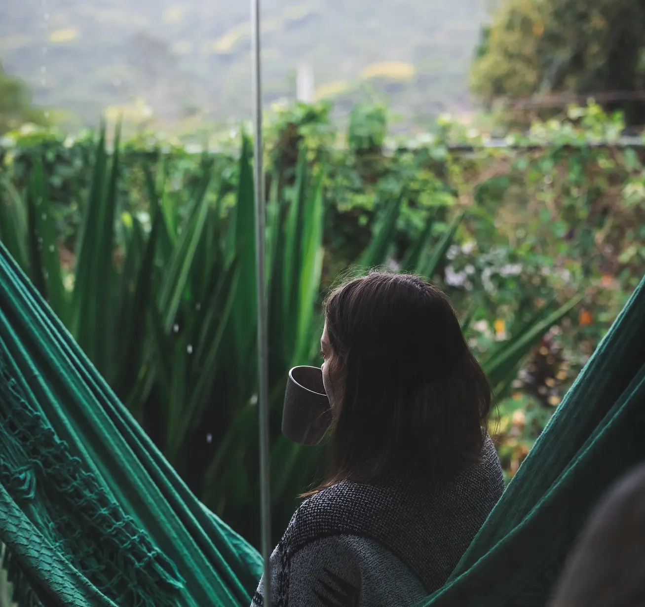 Daniella sitting in a green hammock outdoors, drinking from a gray cup, with green plants and mountains in the background.