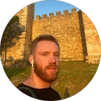 Bearded man wearing wireless earphones outdoors with a stone fortress wall in the background during sunset.