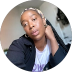Portrait of a woman with a headscarf and braided hair, resting her hand on her neck, wearing a dark top and a white shirt.