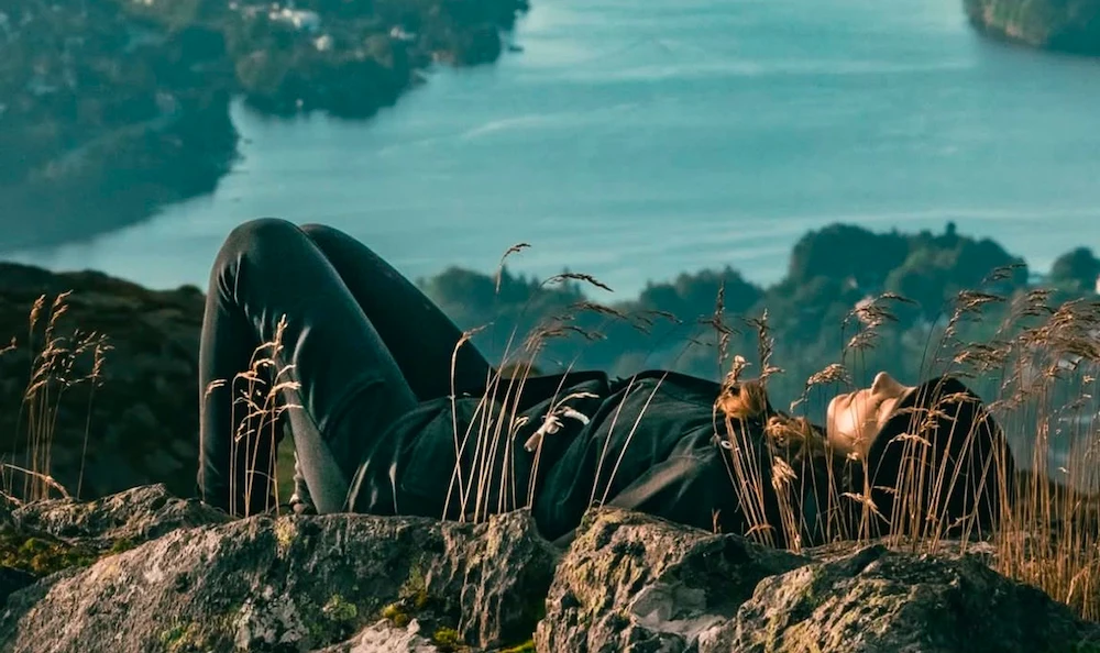 Woman wearing headphones and an orange dress working on a tablet while taking notes at a wooden table with two cups.