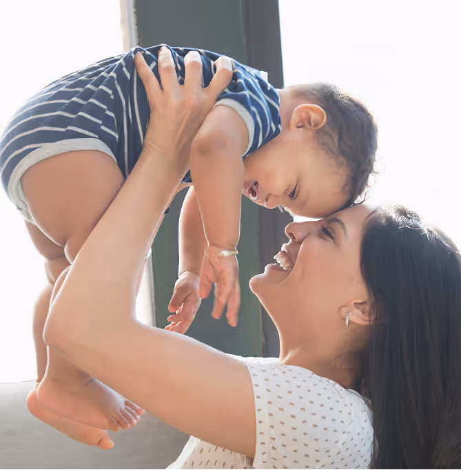 A photograph of a mother playing with a baby by carrying the baby over her head and both sharing a cheerful moment