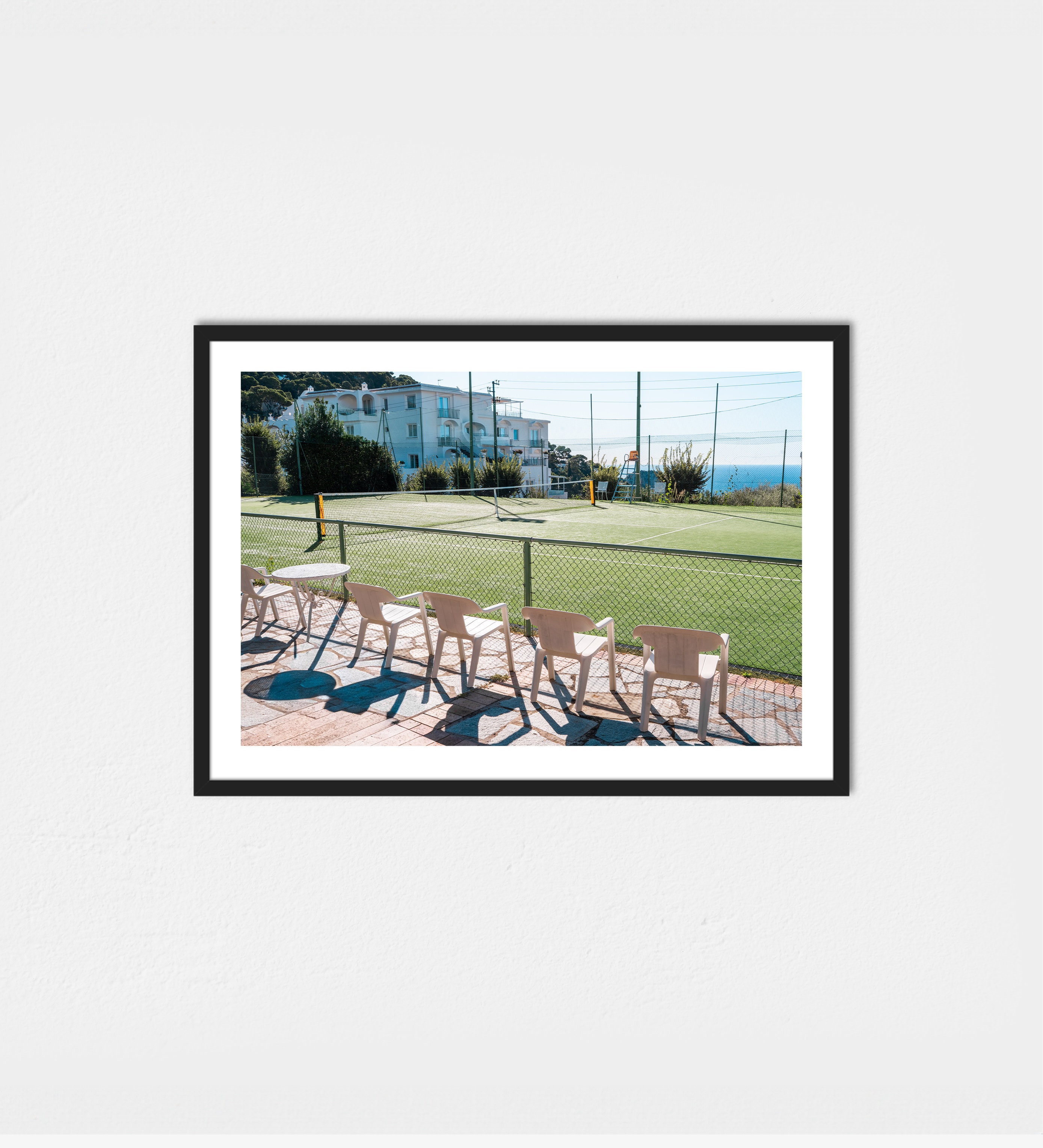 A sunlit tennis court at Hotel Quisisana in Capri, pink chairs casting shadows beside the green court with the Mediterranean beyond.