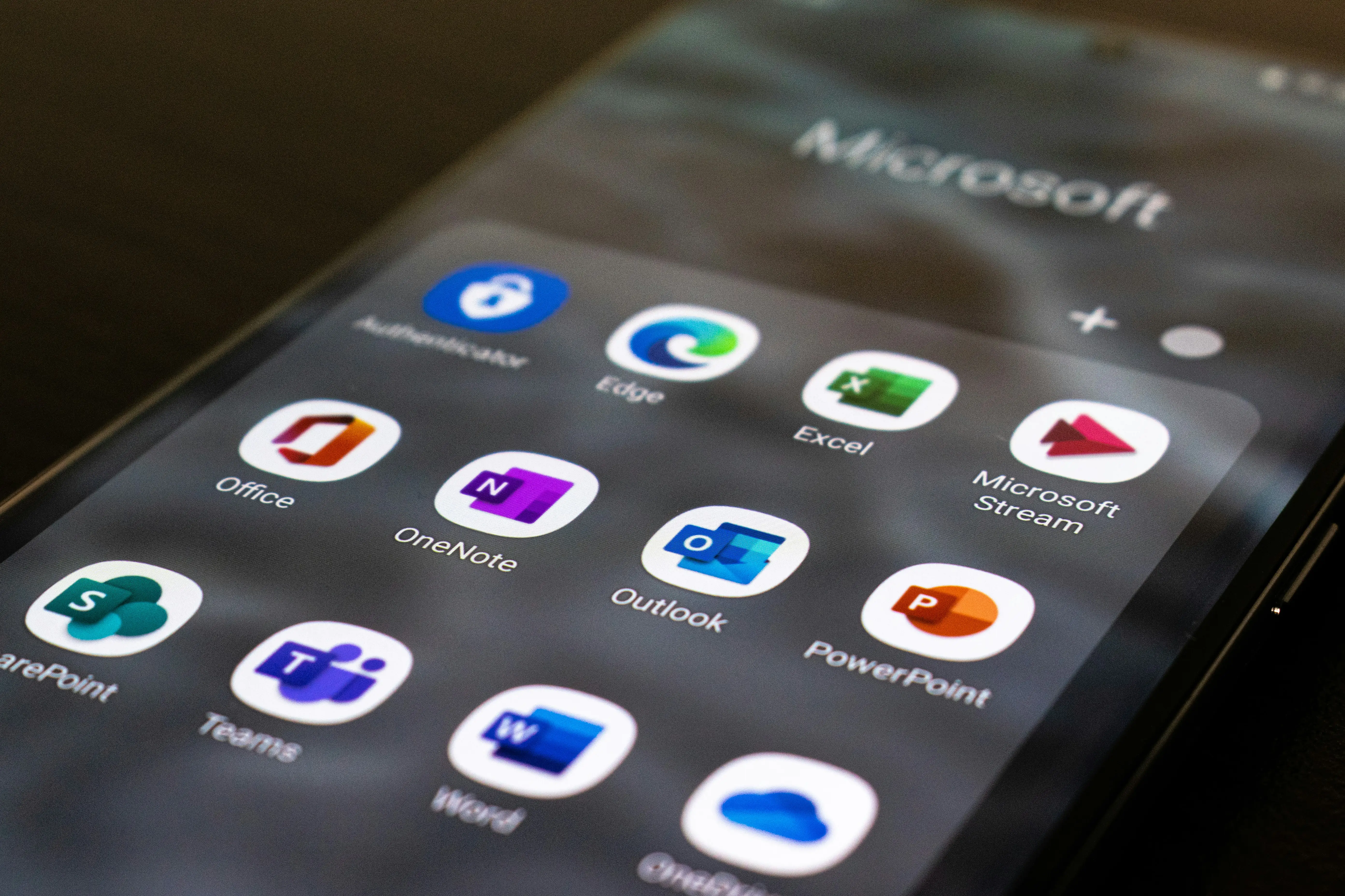 An iPhone opened to a folder of Microsoft apps, like Authenticator, Teams, OpenNote, PowerPoint, Excel, and SharePoint.