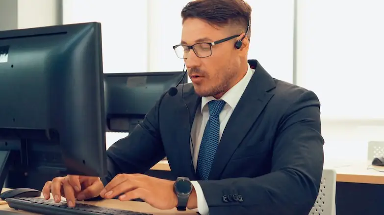 A white man in a suit sits at a computer wearing a headset with a microphone.