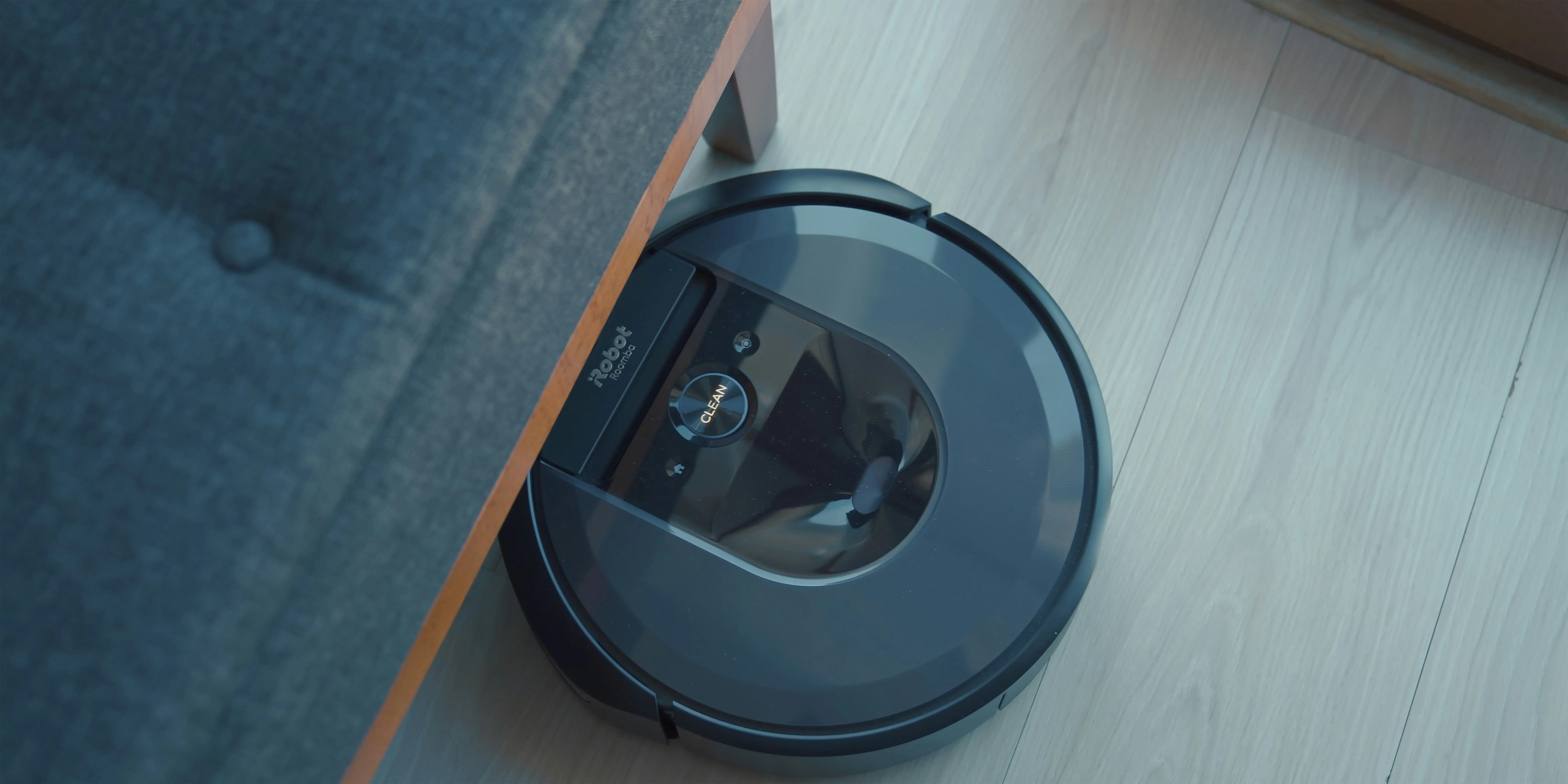 A black robot vacuum partially hidden under a grey piece of furniture.