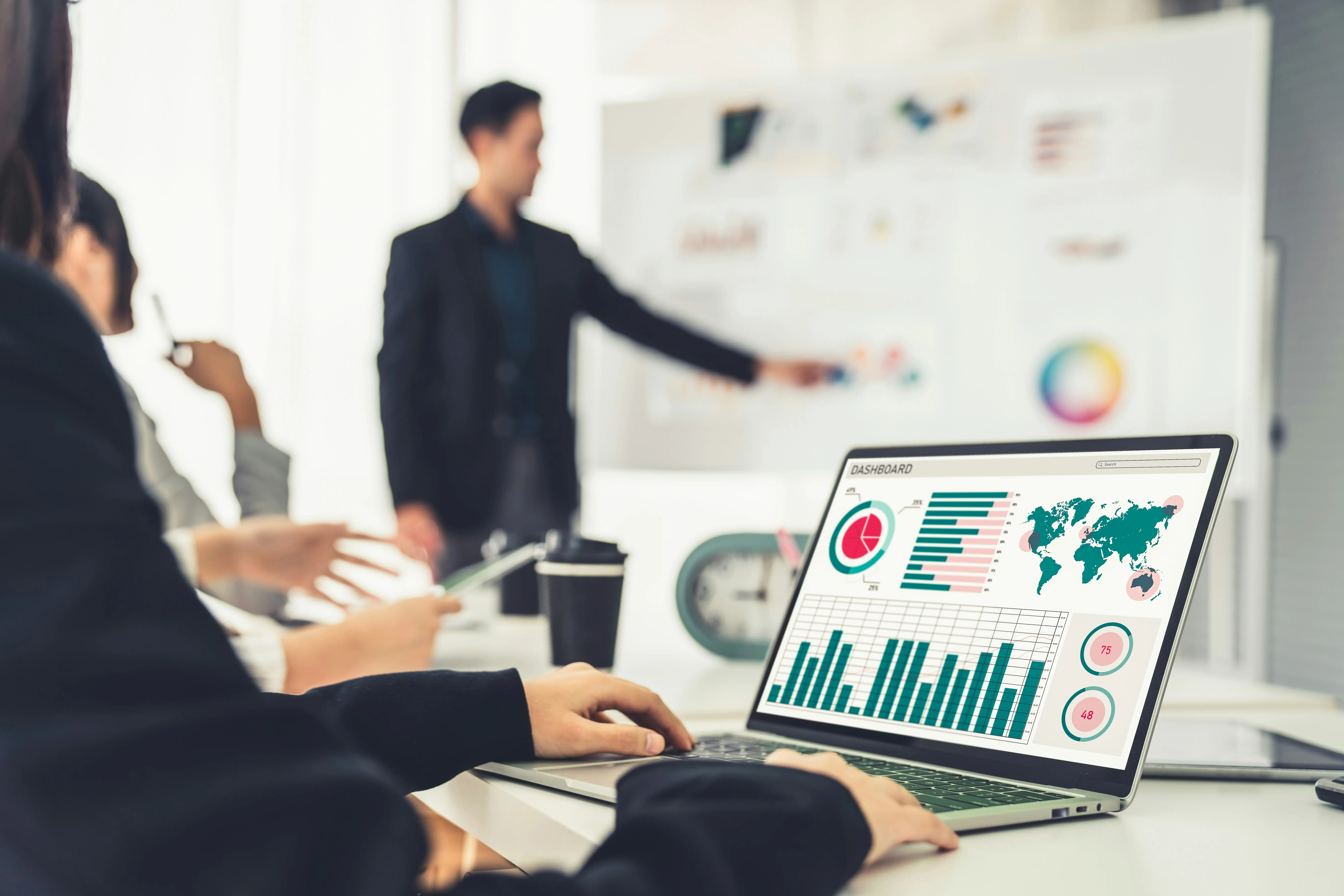 Close-up of a computer with graphs and a map of the world, with a man presenting in the background.