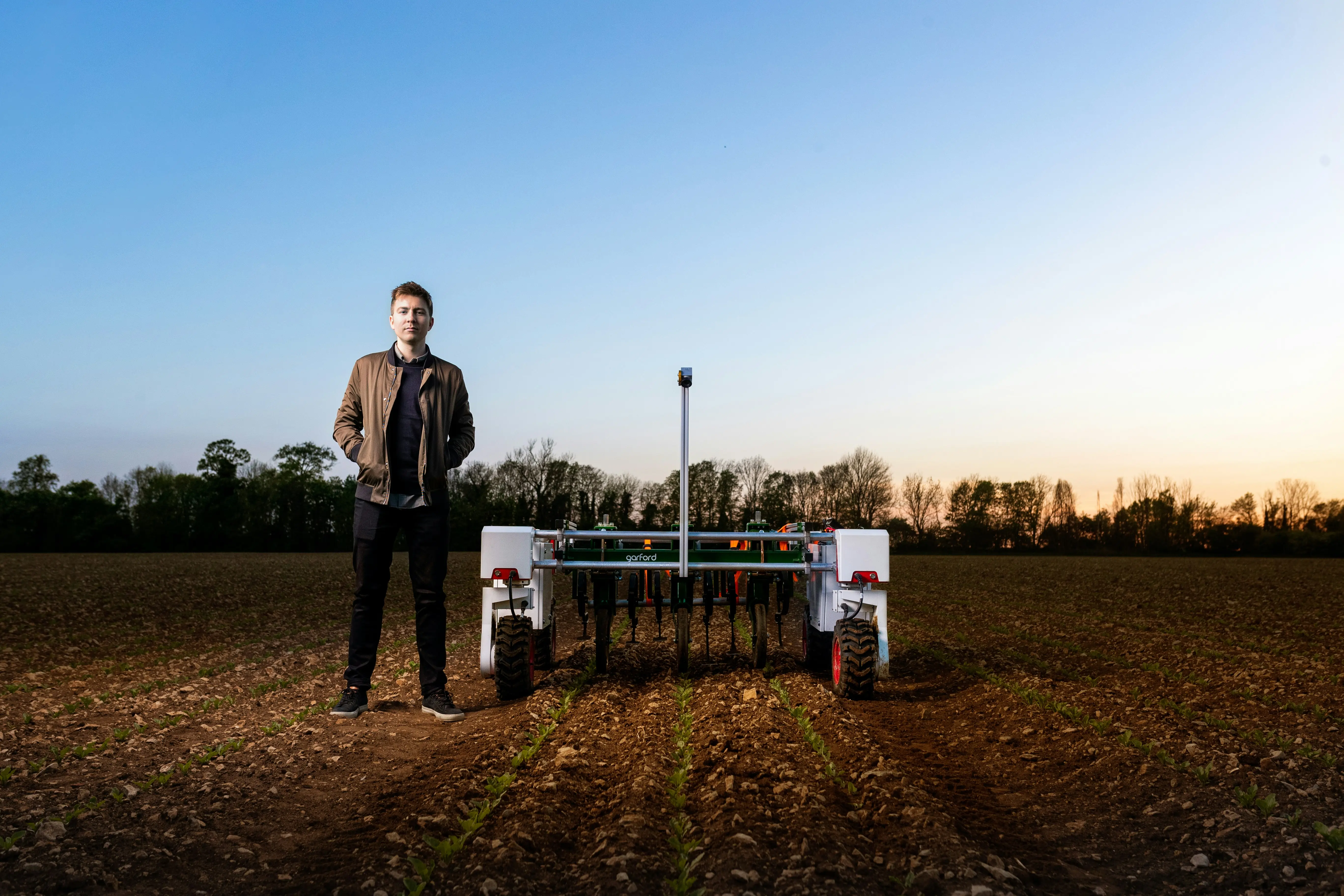 A young, white man stands next to a piece of lean farming machinery on a plowed field at dawn.