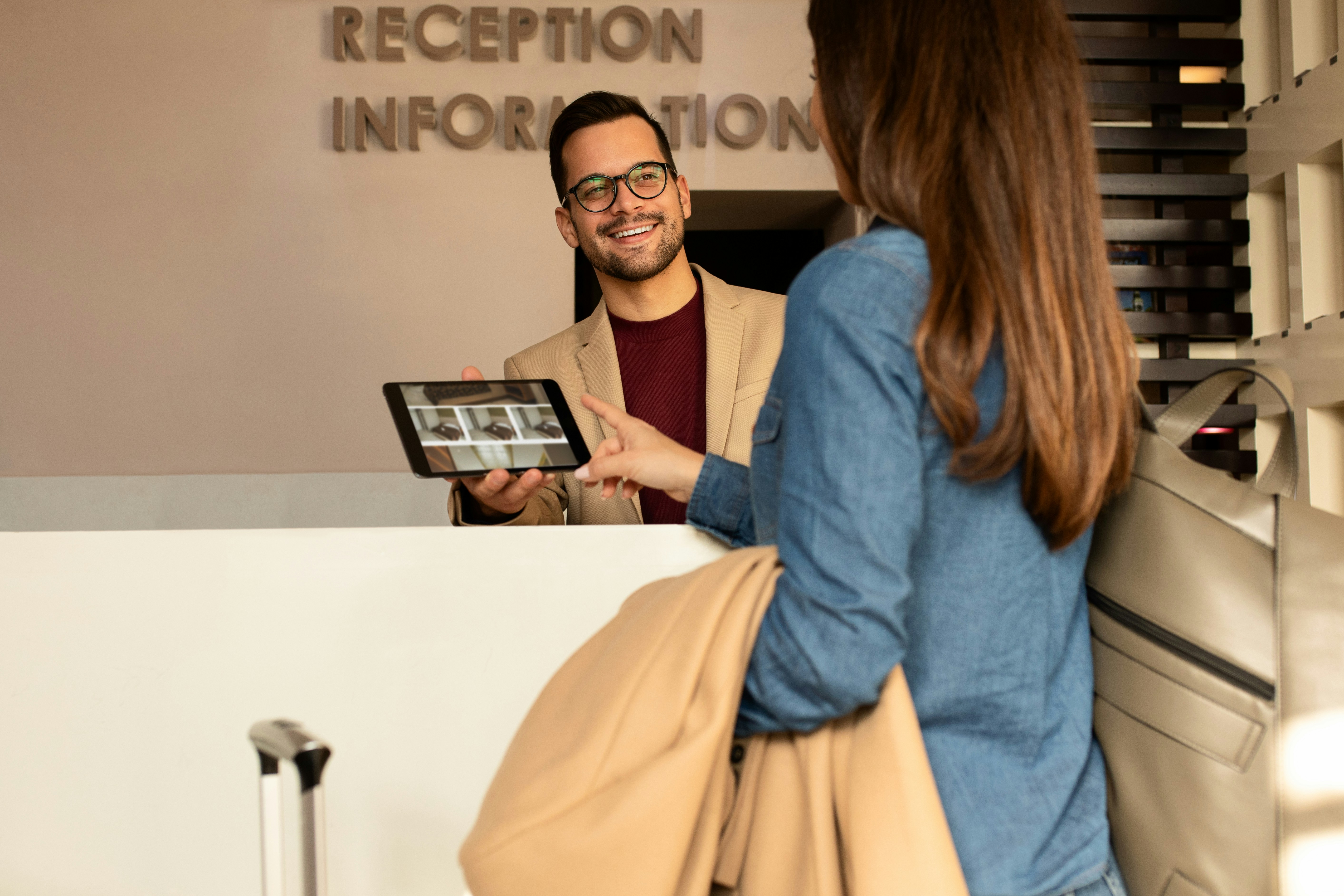 A long-haired woman taps a tablet held by a smiling man behind a desk labeled 'Reception Information'.