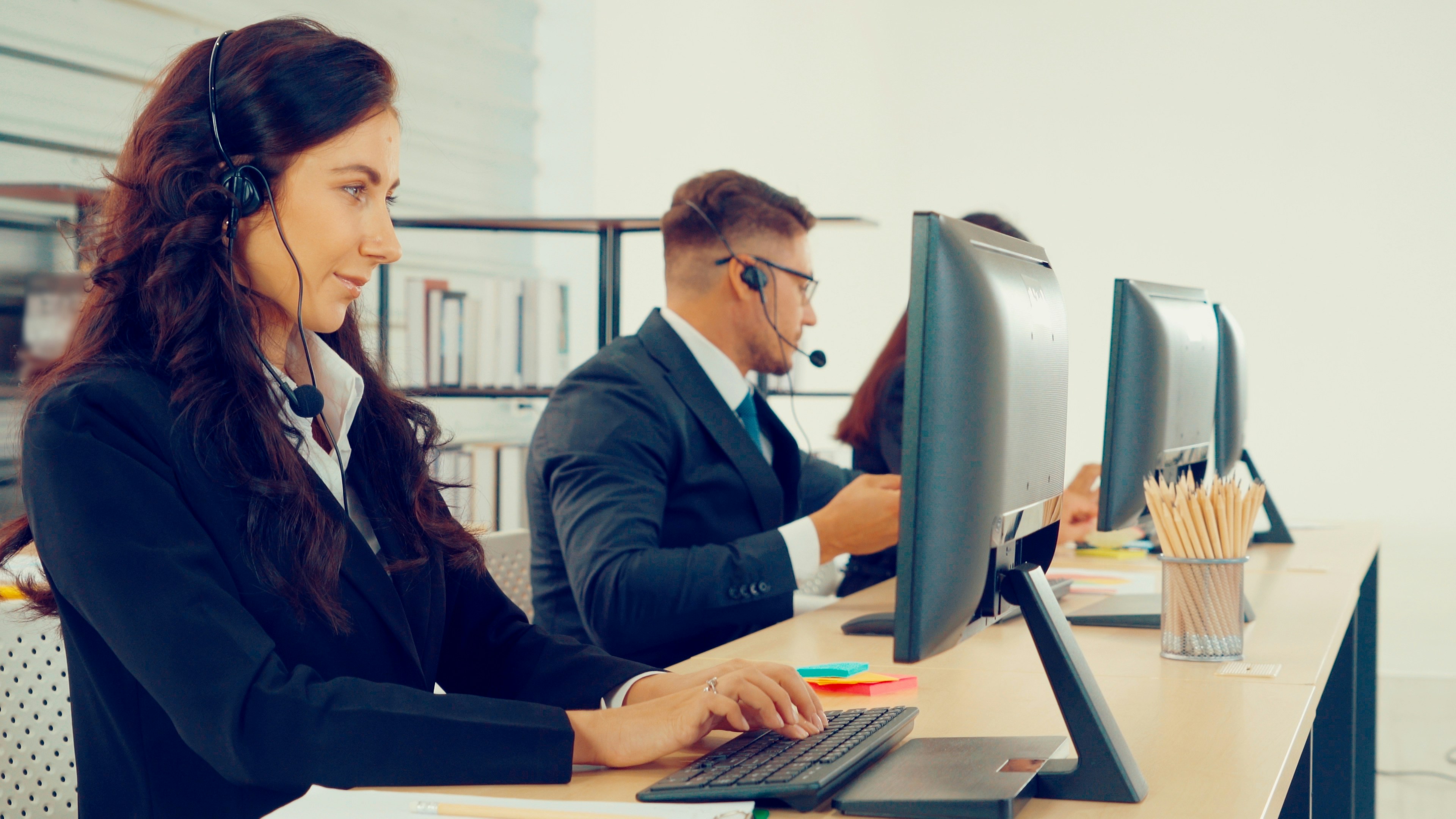 Three employees in suits sit at computers wearing headsets.