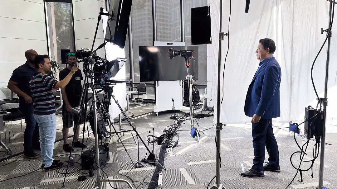 Man in a blue suit standing in front of a camera and professional lighting setup with a crew adjusting equipment in a modern office space.