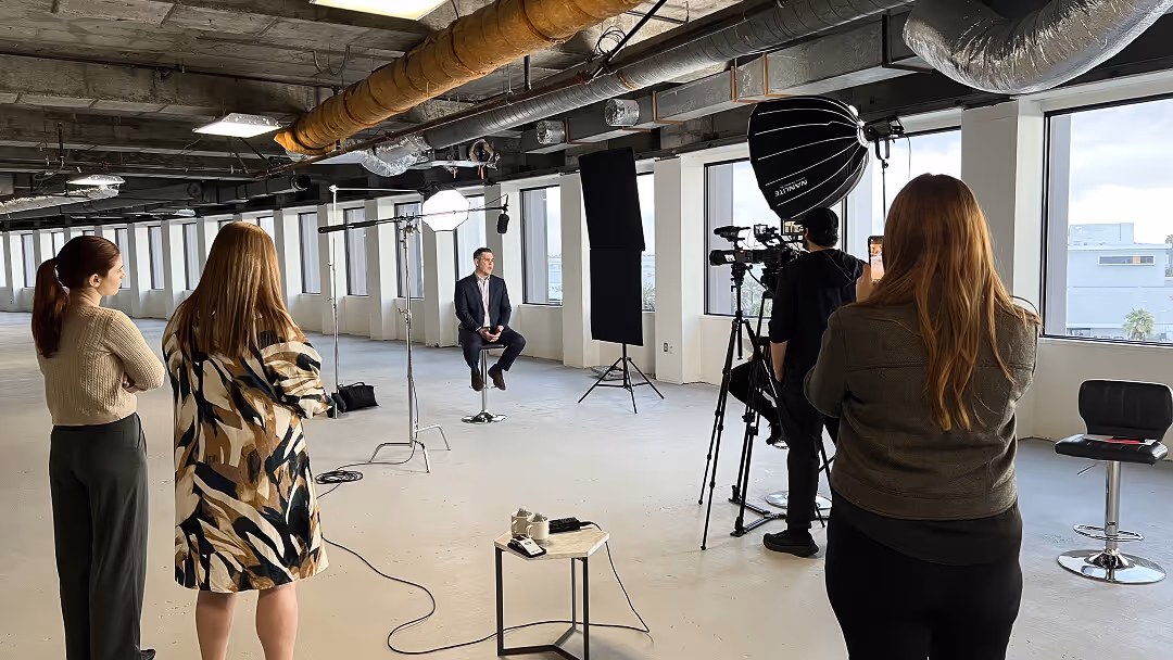 Man in suit sitting on stool in an industrial studio while crew films and lights him with professional equipment.