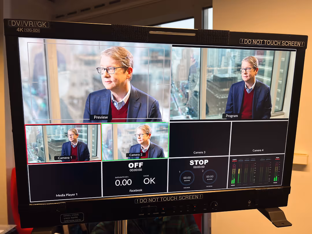 Video production monitor displaying multiple camera feeds of a man in glasses and a navy blazer with a cityscape background.