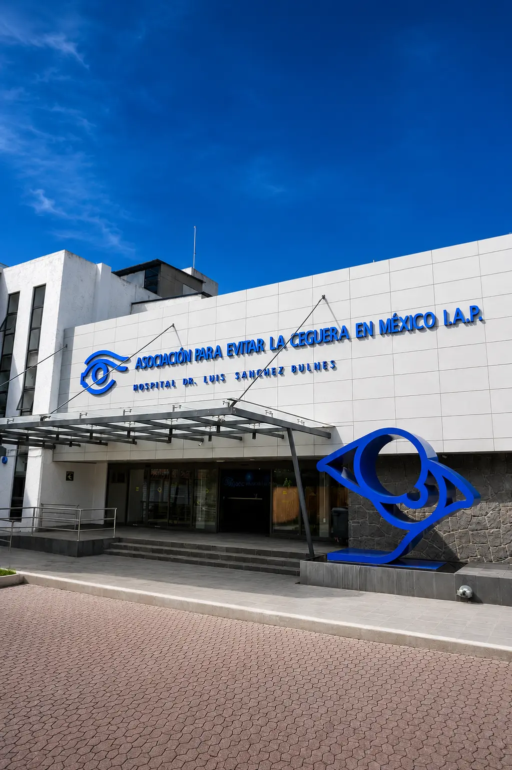 Fachada principal del Hospital Dr. Luis Sánchez Bulnes de APEC, con señalización azul, escultura de un ojo en la entrada y cielo azul despejado.