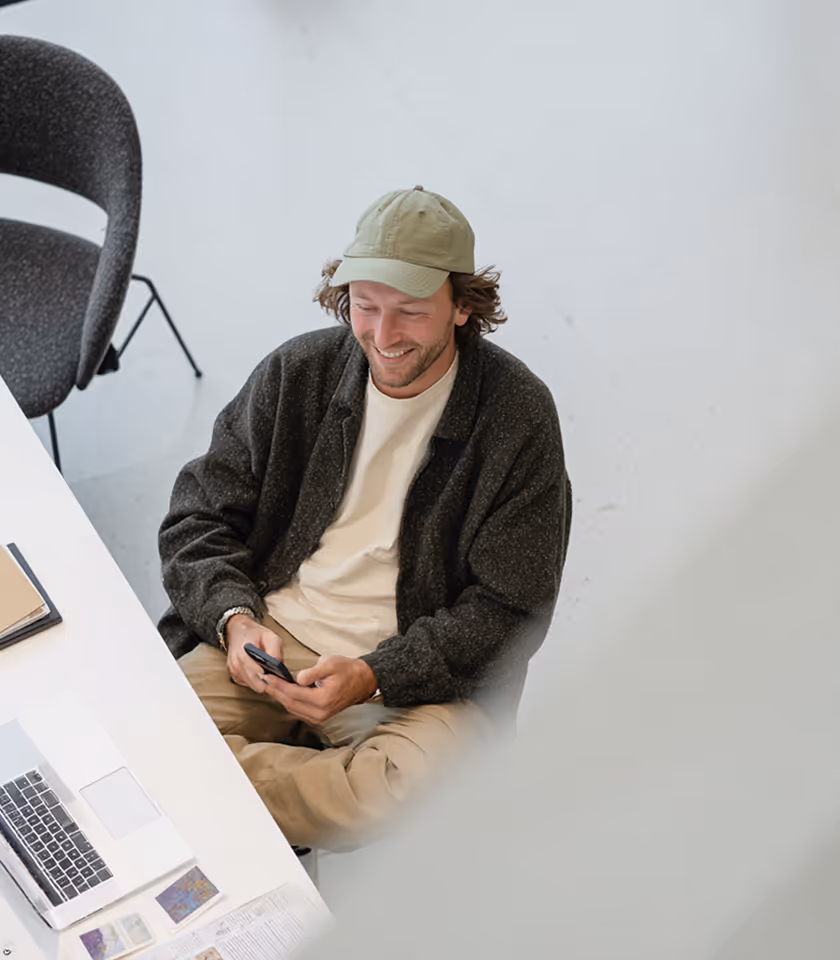 Smiling man in a green cap and dark jacket sitting at a white desk looking at his phone.