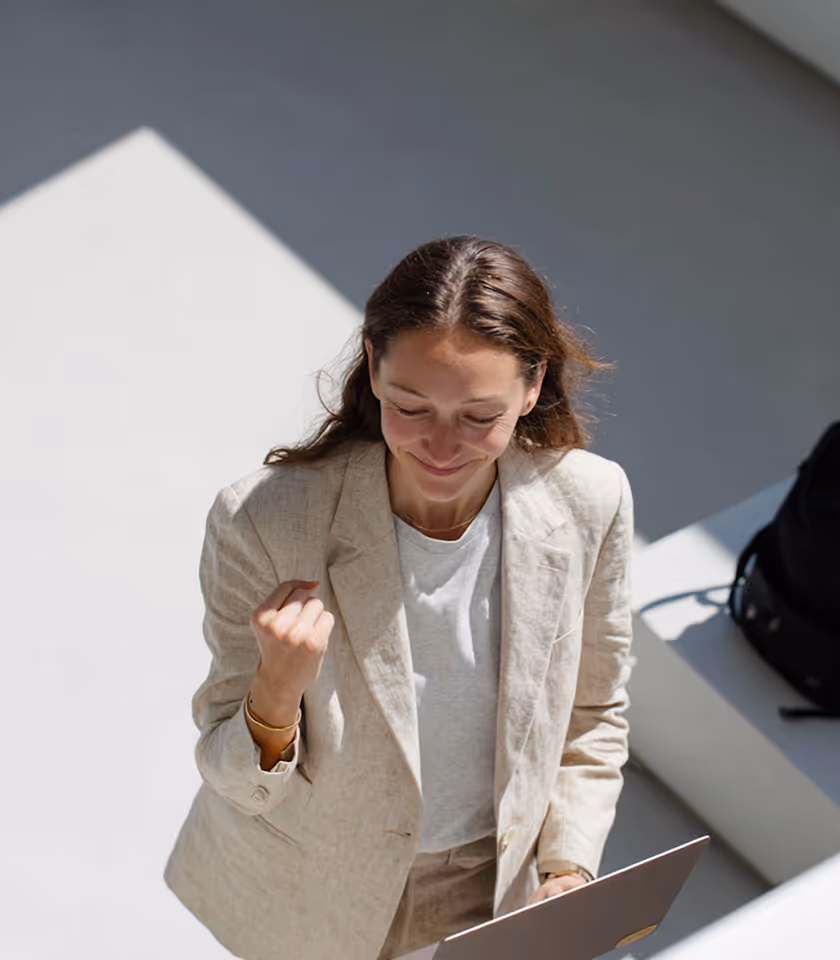 Smiling woman in beige blazer looking at a laptop and making a fist of success.