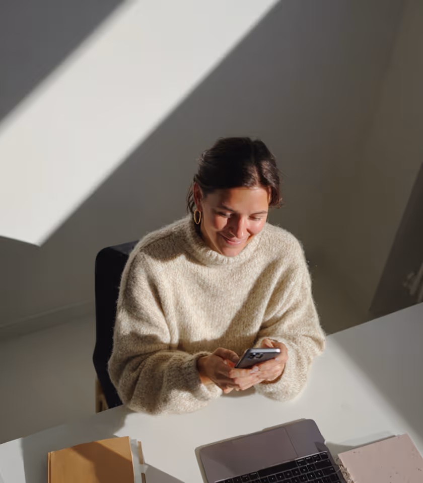 Woman in a beige sweater smiling while looking at her smartphone at a white desk with an open laptop and notebook.