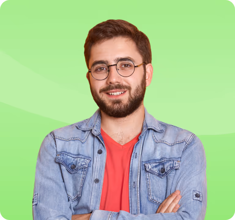 Smiling man with beard and round glasses wearing a red shirt and light denim jacket standing with arms crossed against a green background.