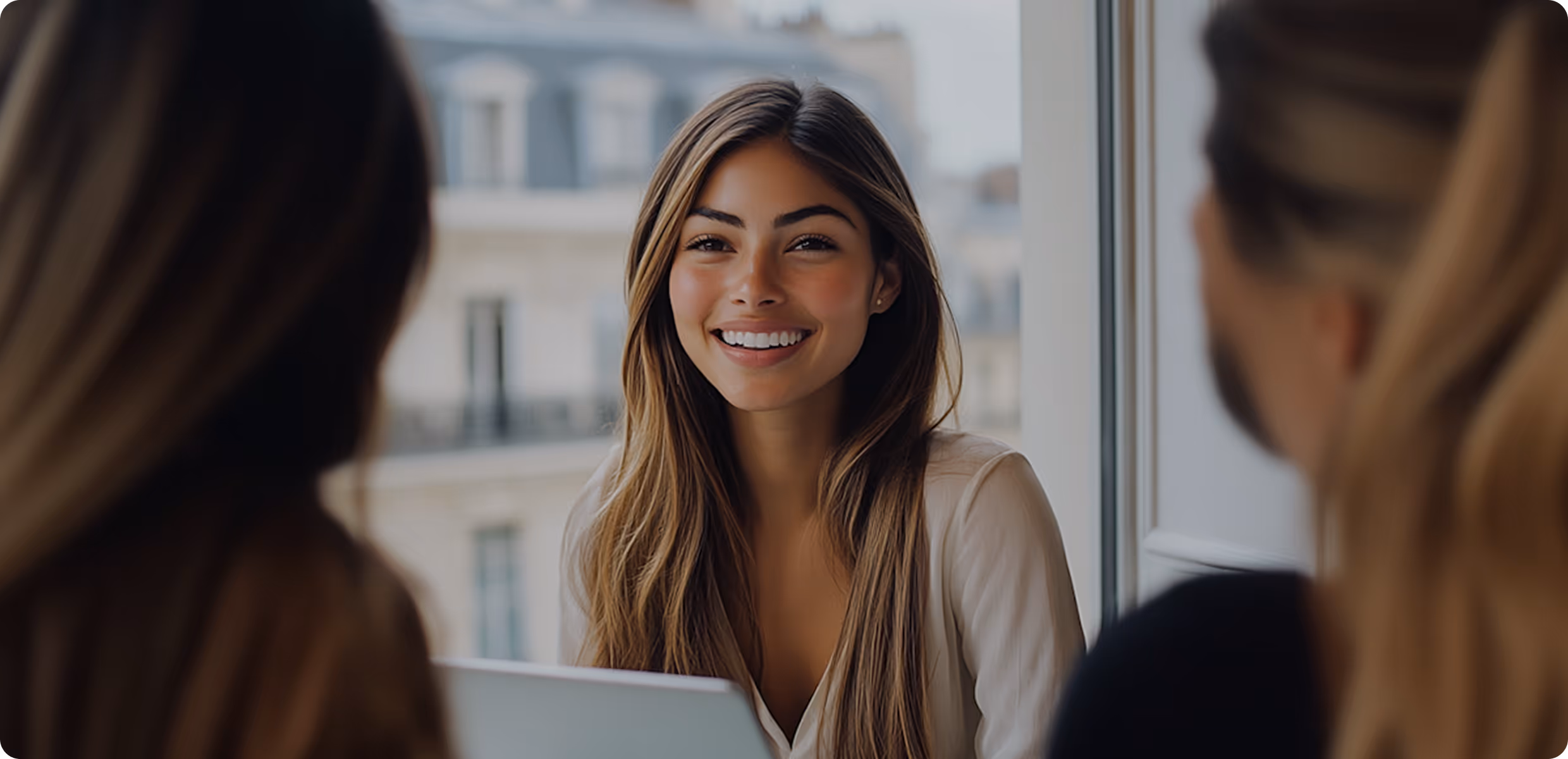 Smiling woman with long hair sitting indoors, facing two other people blurred in the foreground.