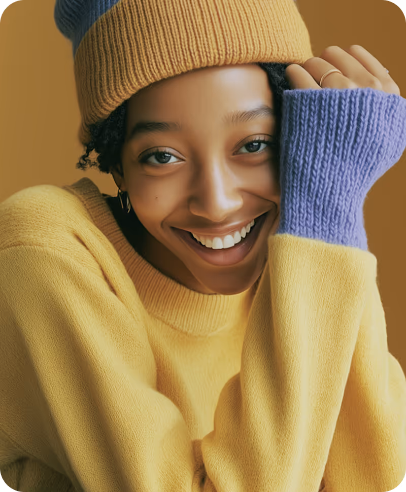 Smiling young woman wearing a yellow sweater, color-block blue and yellow knitted gloves, and a matching beanie against a warm brown background.