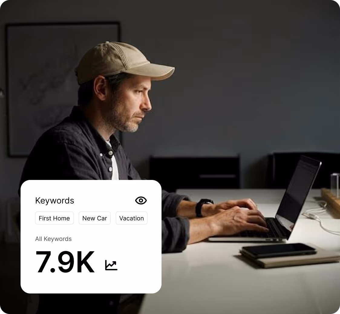 Man wearing a beige cap and black shirt typing on a laptop at a desk with a smartphone and notebook nearby.