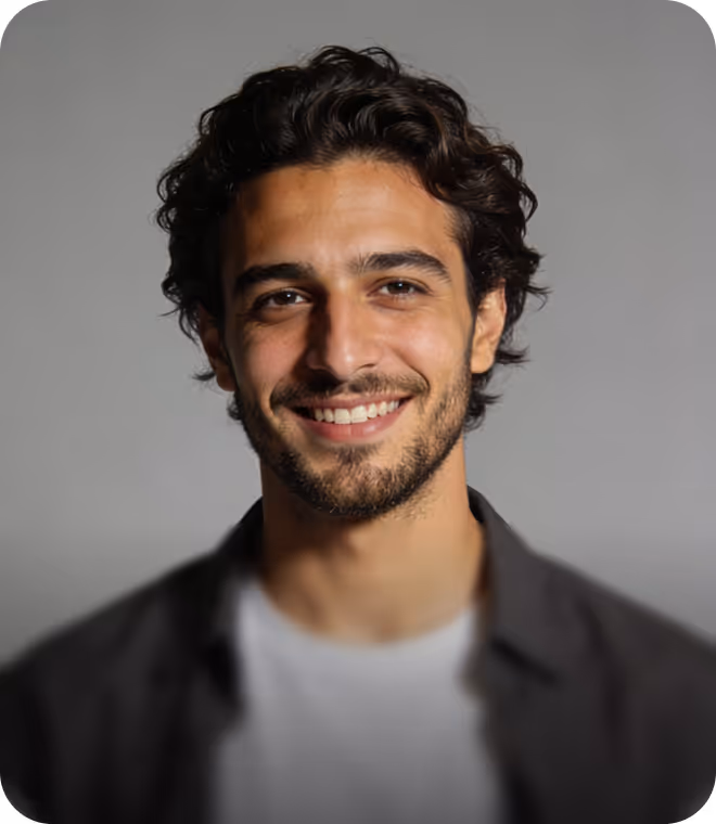 Smiling young man with curly dark hair and facial hair wearing a gray jacket over a white shirt against a gray background.