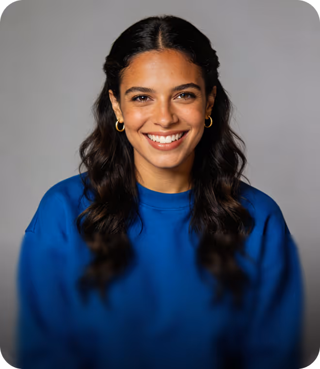 Smiling woman with long dark wavy hair wearing gold hoop earrings and a blue sweater against a gray background.