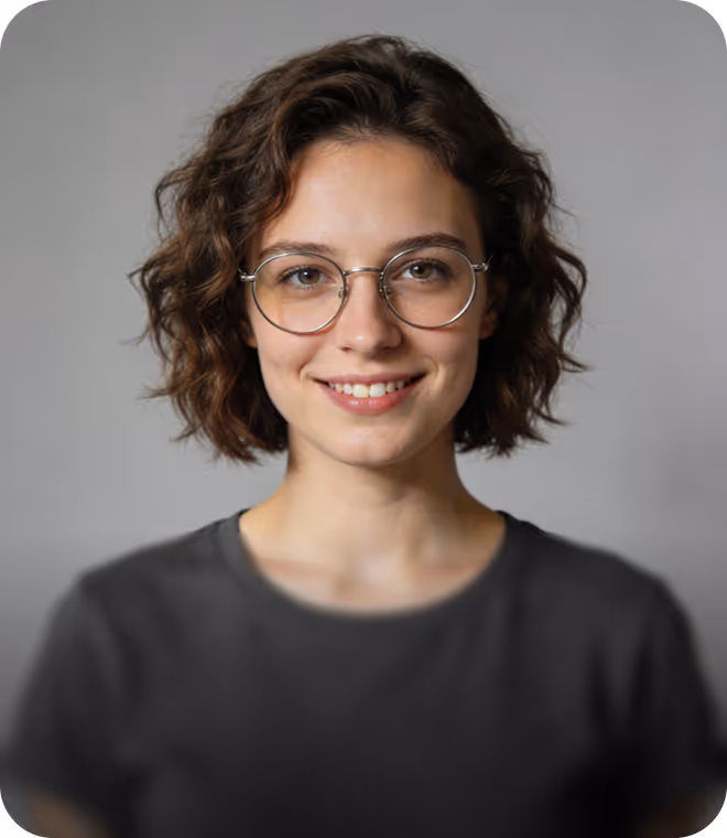 Smiling young woman with short curly brown hair and round glasses wearing a dark gray shirt against a plain gray background.
