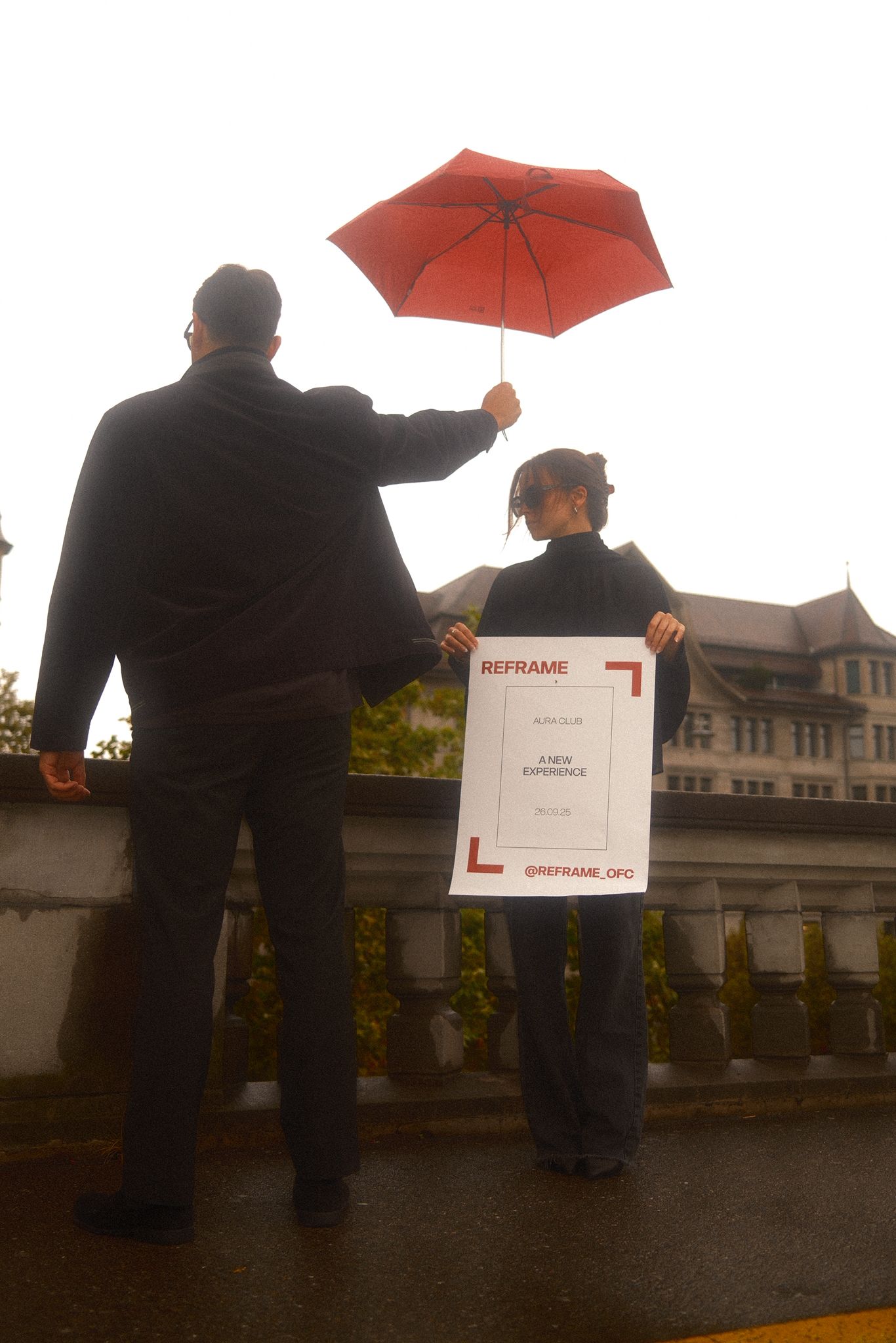 Zwei Personen auf einer Brücke, eine hält einen roten Regenschirm, die andere ein REFRAME-Plakat für das Event in Zürich.