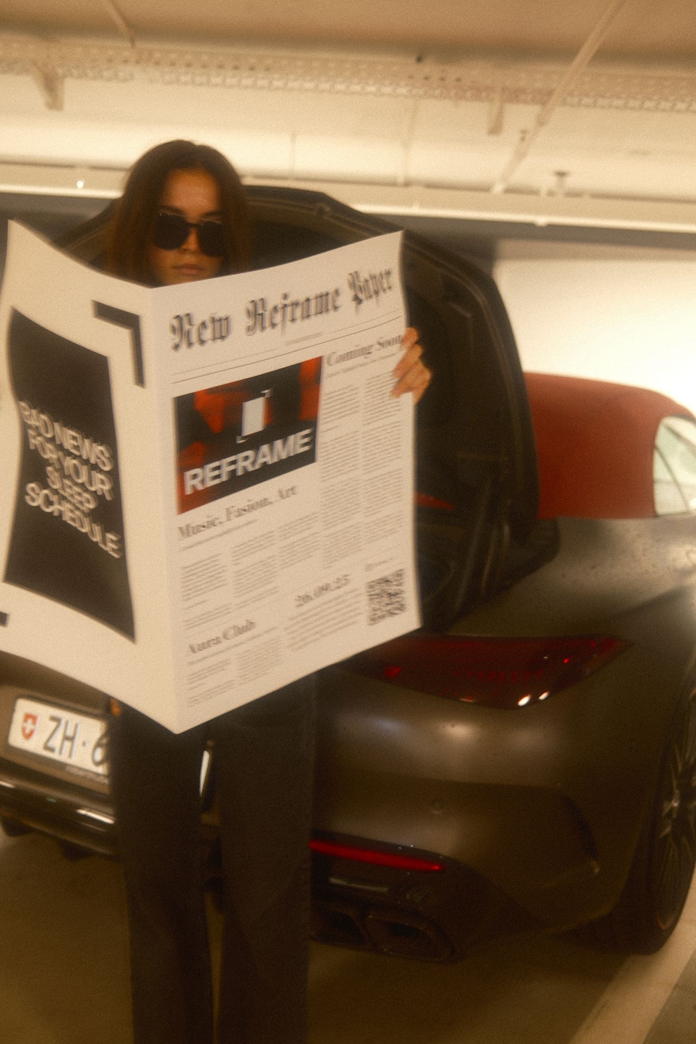 Person mit Sonnenbrille liest REFRAME-Zeitung in einer Tiefgarage vor einem geparkten Auto.