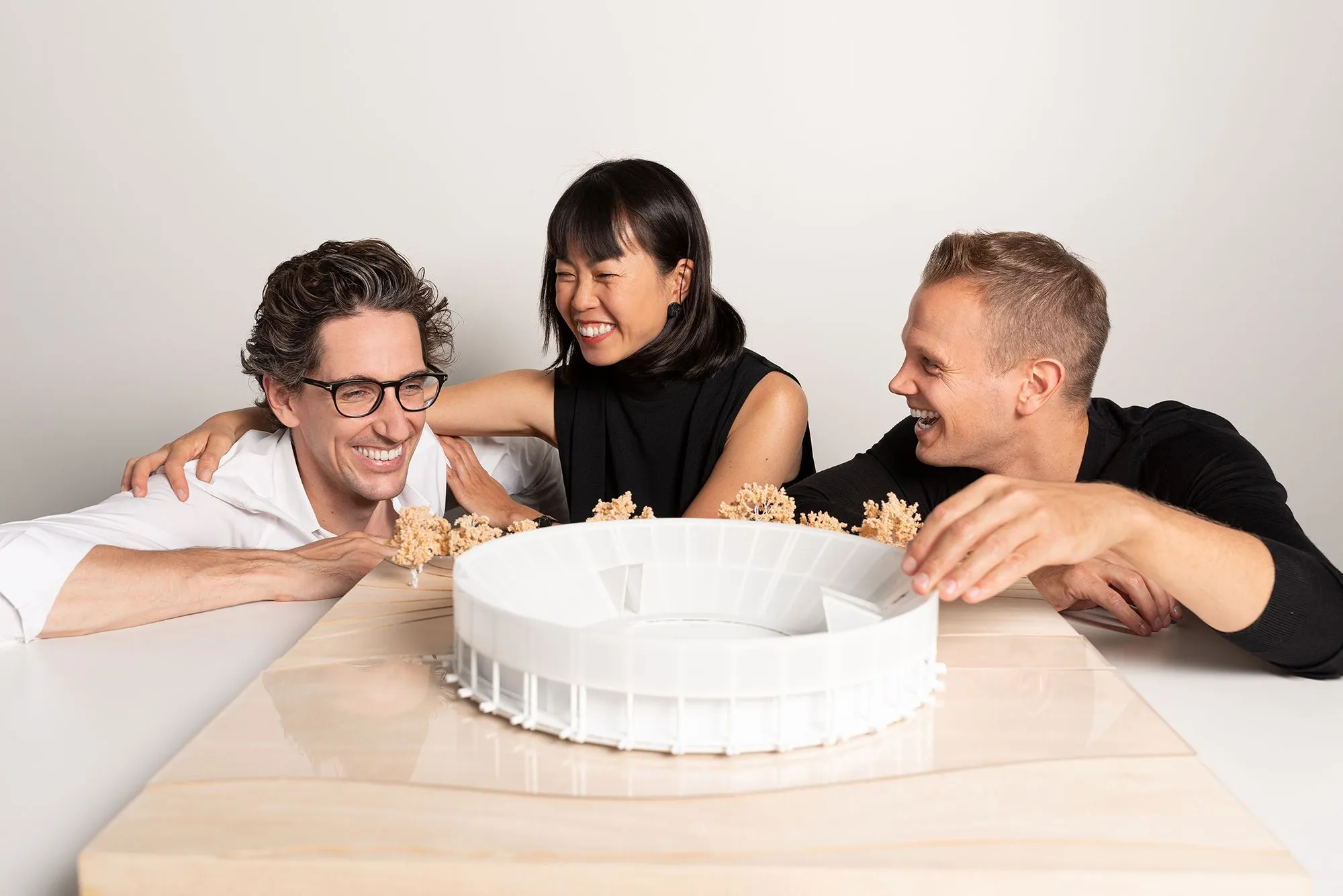 Three architects smiling and discussing a circular building model, symbolizing teamwork and creative collaboration.