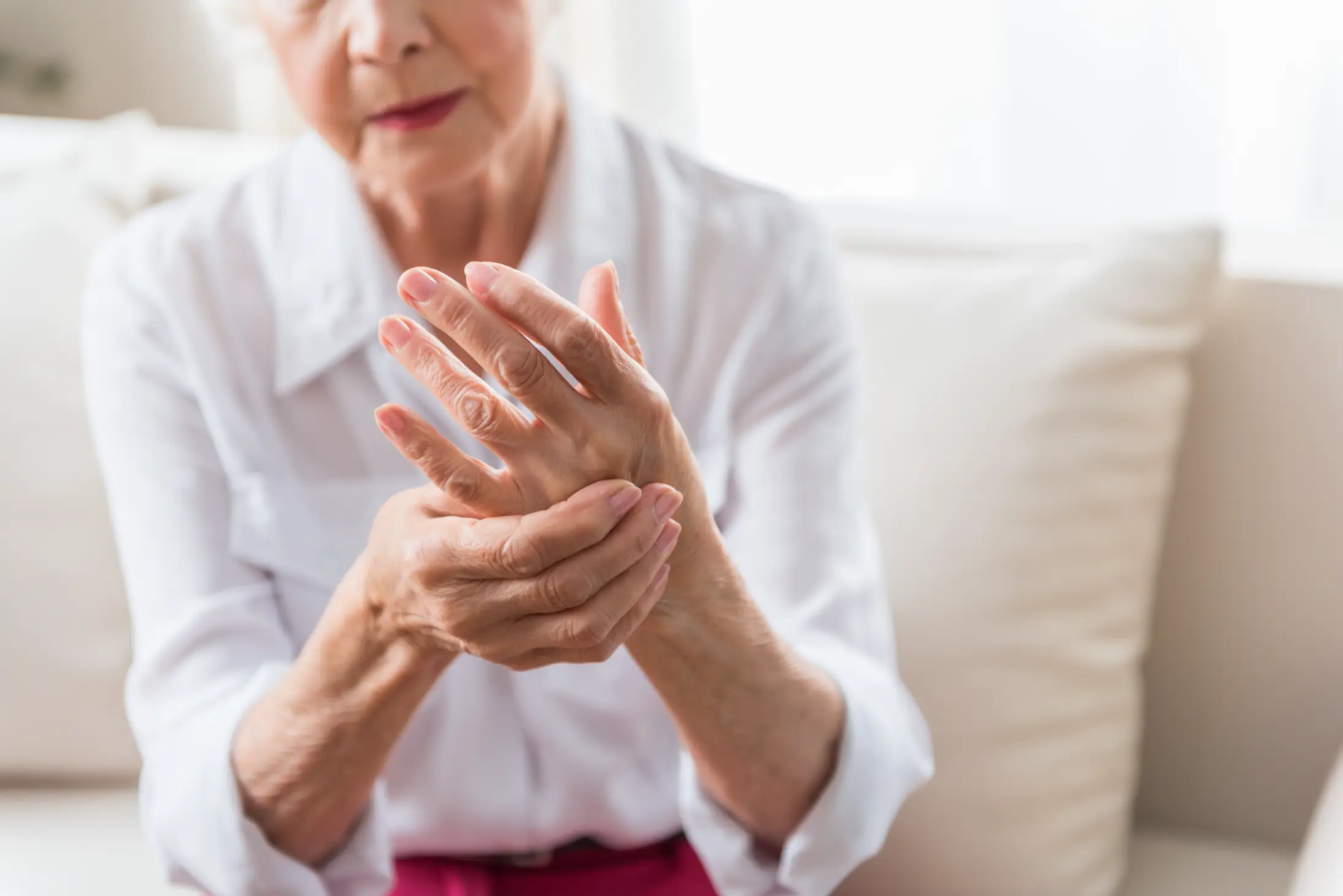 Elderly woman in white clothing grimacing as she massages her hand, indicating joint pain or arthritis.