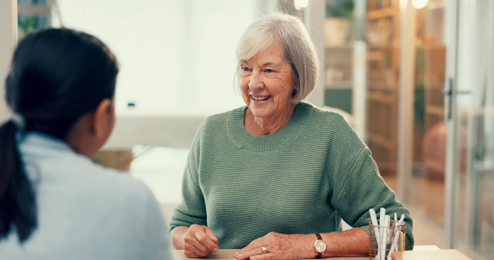 A senior woman with short gray hair, wearing a green sweater, is laughing and talking to someone across from her. The setting looks comfortable and conversational, possibly a consultation for this chiropractic clinic.