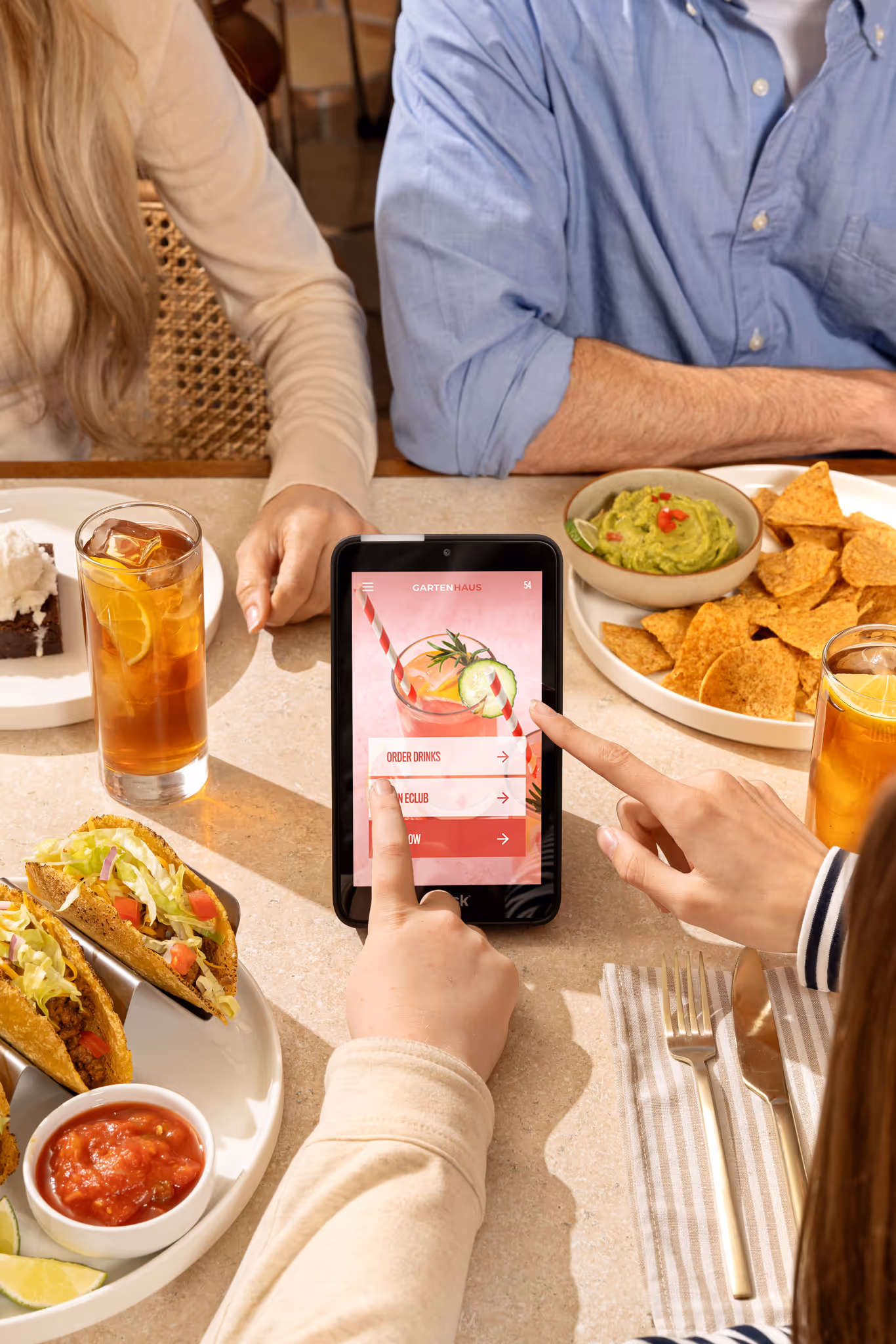 Two people sitting at a table with tacos, chips and guacamole, and iced tea, using a digital menu on a tablet to order drinks.