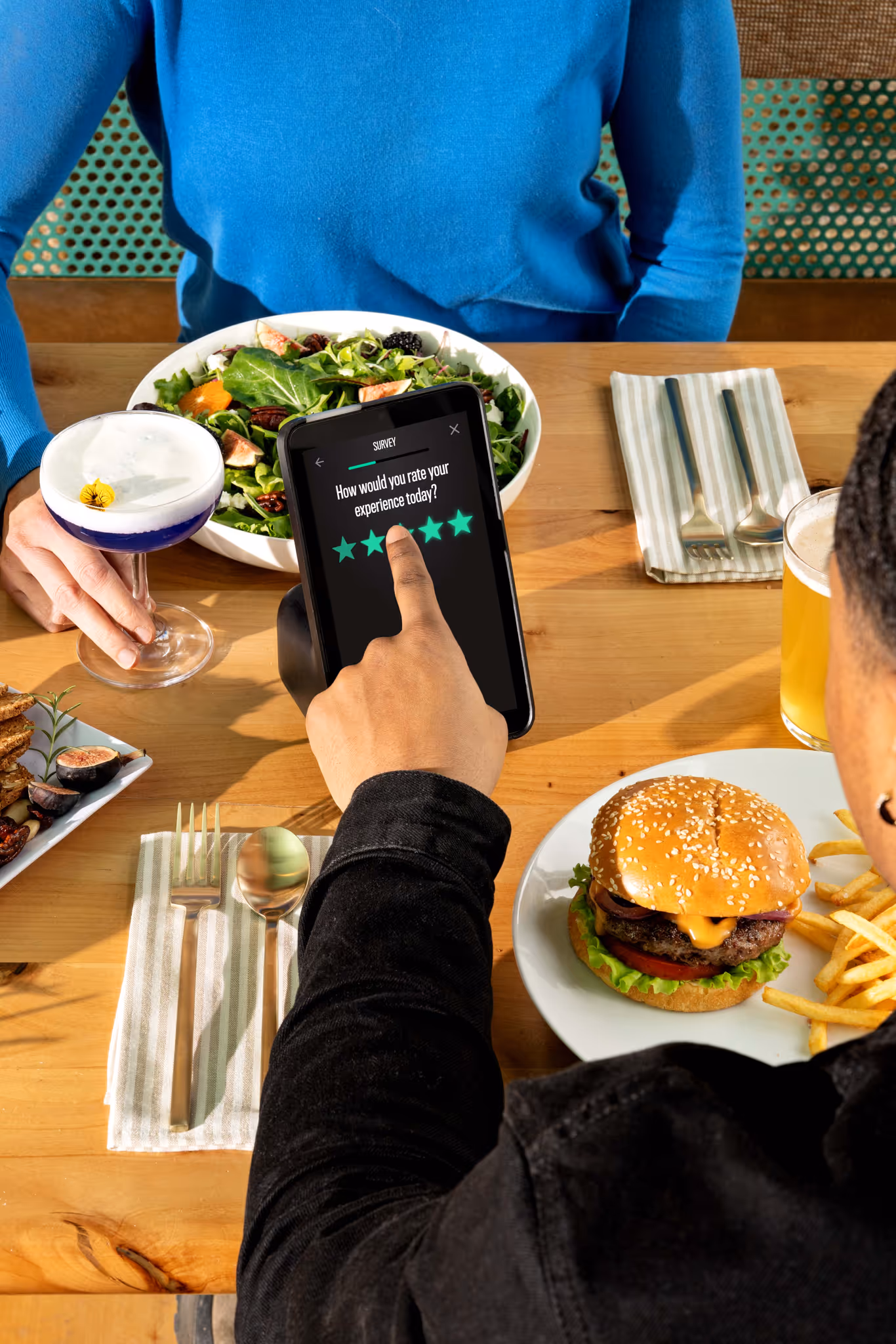 Person rating their dining experience on a tablet showing a five-star survey while sitting at a table with a burger, fries, salad, and drinks.