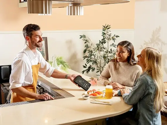 Barista handing a payment device to two women sitting at a bar counter with drinks and fruit on the countertop.