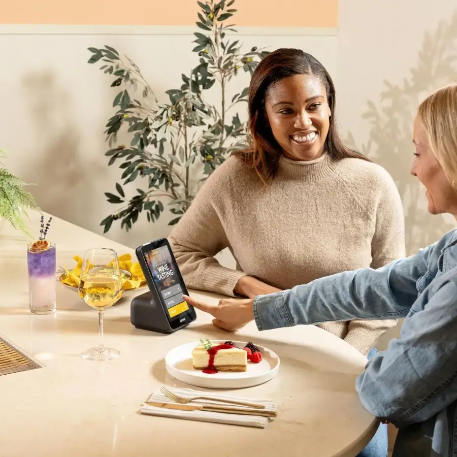 Two women sitting at a table with drinks and cheesecake, smiling and interacting with a small touchscreen device.