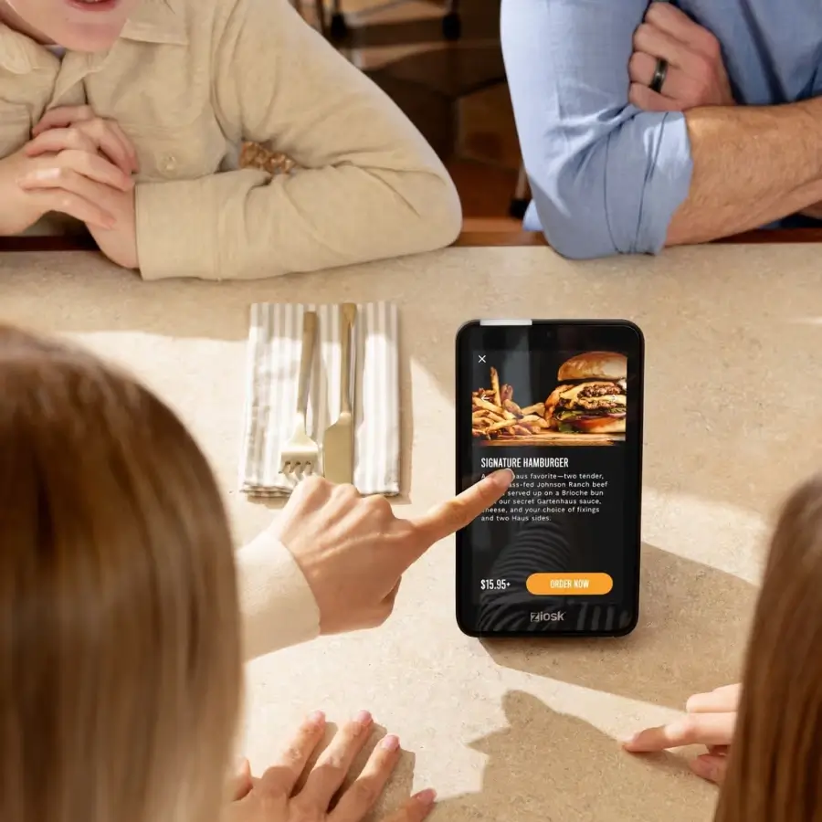 Four people sitting at a table looking at a digital menu on a tablet featuring a signature hamburger with fries and an order button.
